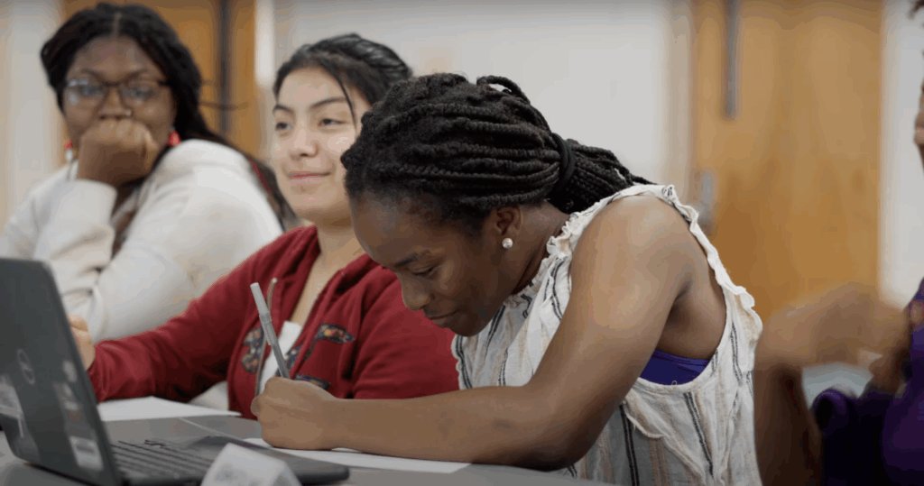 Students seated at a table take notes and use a laptop.